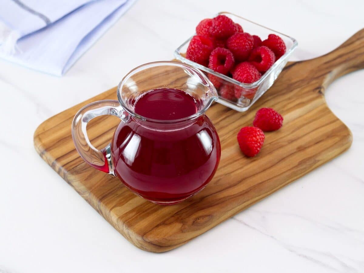 Angled horizontal shot of a glass pitcher filled with bright red raspberry syrup next to a bowl of fresh raspberries on top of a wooden cutting board.