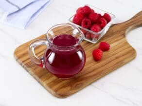 Angled horizontal shot of a glass pitcher filled with bright red raspberry syrup next to a bowl of fresh raspberries on top of a wooden cutting board.