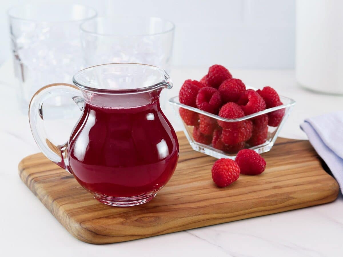 Angled horizontal shot of a glass pitcher filled with bright red homemade raspberry syrup next to a bowl of fresh raspberries on top of a wooden cutting board.