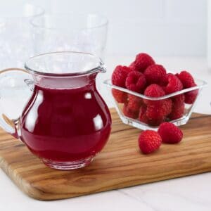Angled horizontal shot of a glass pitcher filled with bright red homemade raspberry syrup next to a bowl of fresh raspberries on top of a wooden cutting board.