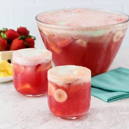 Horizontal shot of a punch bowl filled with a red foamy beverage. Two glasses of the punch sit next to the bowl next to a dish of fresh berries.