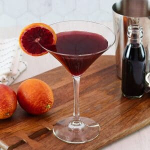 Horizontal image of a martini glass filled with a dark red cocktail and garnished with a slice of blood orange. Two blood oranges and a steel cocktail jigger sit off to the sides.