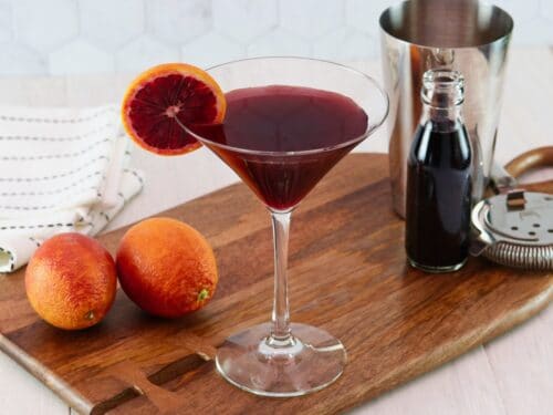 Horizontal image of a martini glass filled with a dark red cocktail and garnished with a slice of blood orange. Two blood oranges and a steel cocktail jigger sit off to the sides.