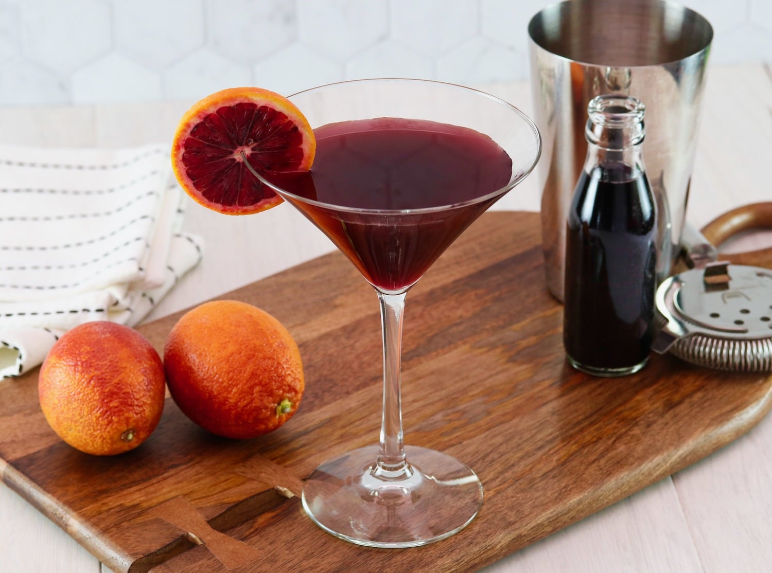 Horizontal image of a martini glass filled with a dark red cocktail and garnished with a slice of blood orange. Two blood oranges and a steel cocktail jigger sit off to the sides.