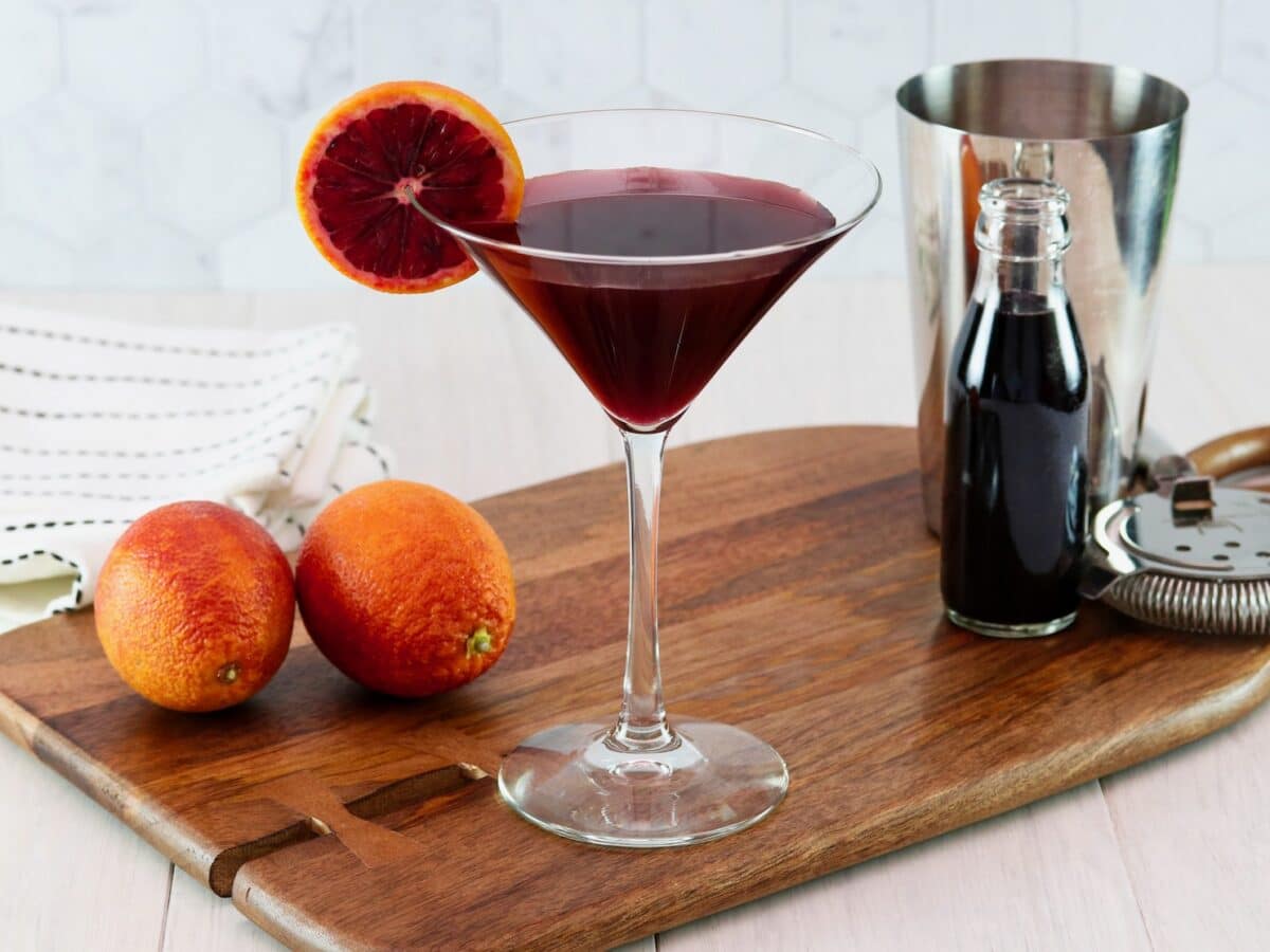Horizontal image of a martini glass filled with a dark red cocktail and garnished with a slice of blood orange. Two blood oranges and a steel cocktail jigger sit off to the sides.