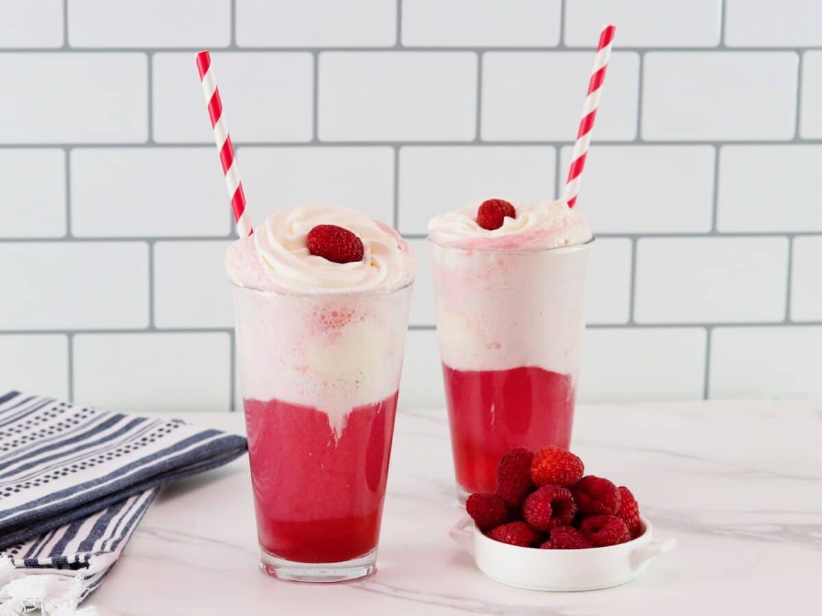 Horizontal shot of two berry ice cream floats in tall glasses. The bright pink colored ice cream soda floats are garnished with whipped cream, a raspberry, and a red and white straw.