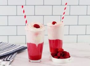 Horizontal shot of two berry ice cream floats in tall glasses. The bright pink colored ice cream soda floats are garnished with whipped cream, a raspberry, and a red and white straw.