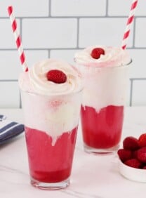 Horizontal shot - two tall glasses of bright pink berry ice cream floats with vanilla ice cream and cream. Soda floats are garnished with whipped cream, a fresh raspberry, and a red and white straw.