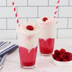 Horizontal shot - two tall glasses of bright pink berry ice cream floats with vanilla ice cream and cream. Soda floats are garnished with whipped cream, a fresh raspberry, and a red and white straw.