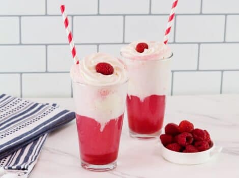 Horizontal shot - two tall glasses of bright pink berry ice cream floats with vanilla ice cream and cream. Soda floats are garnished with whipped cream, a fresh raspberry, and a red and white straw.