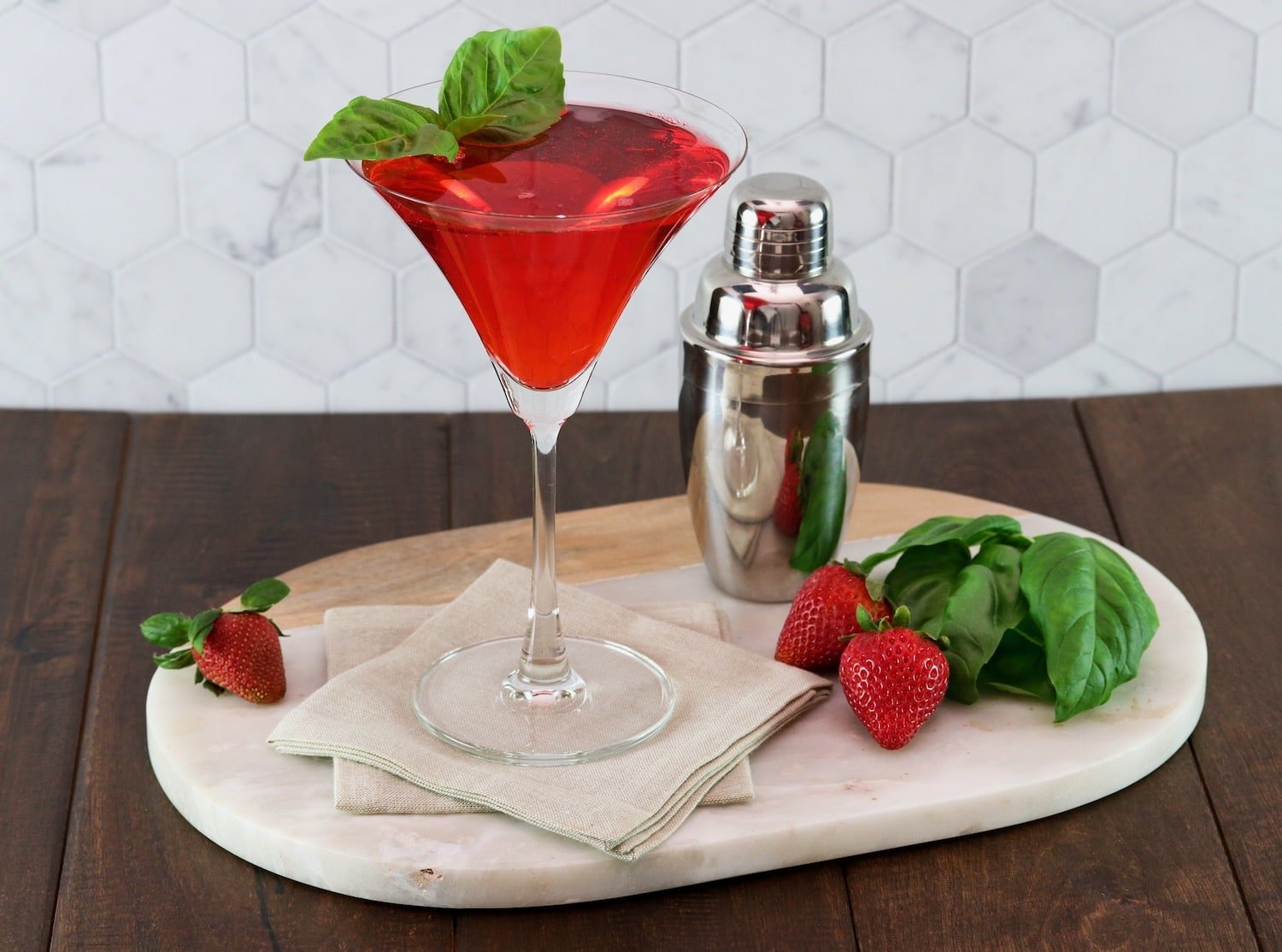 Horizontal shot of a strawberry basil cocktail in martini glass, garnished with a sprig of basil. A steel cocktail shaker sits off to the right side.