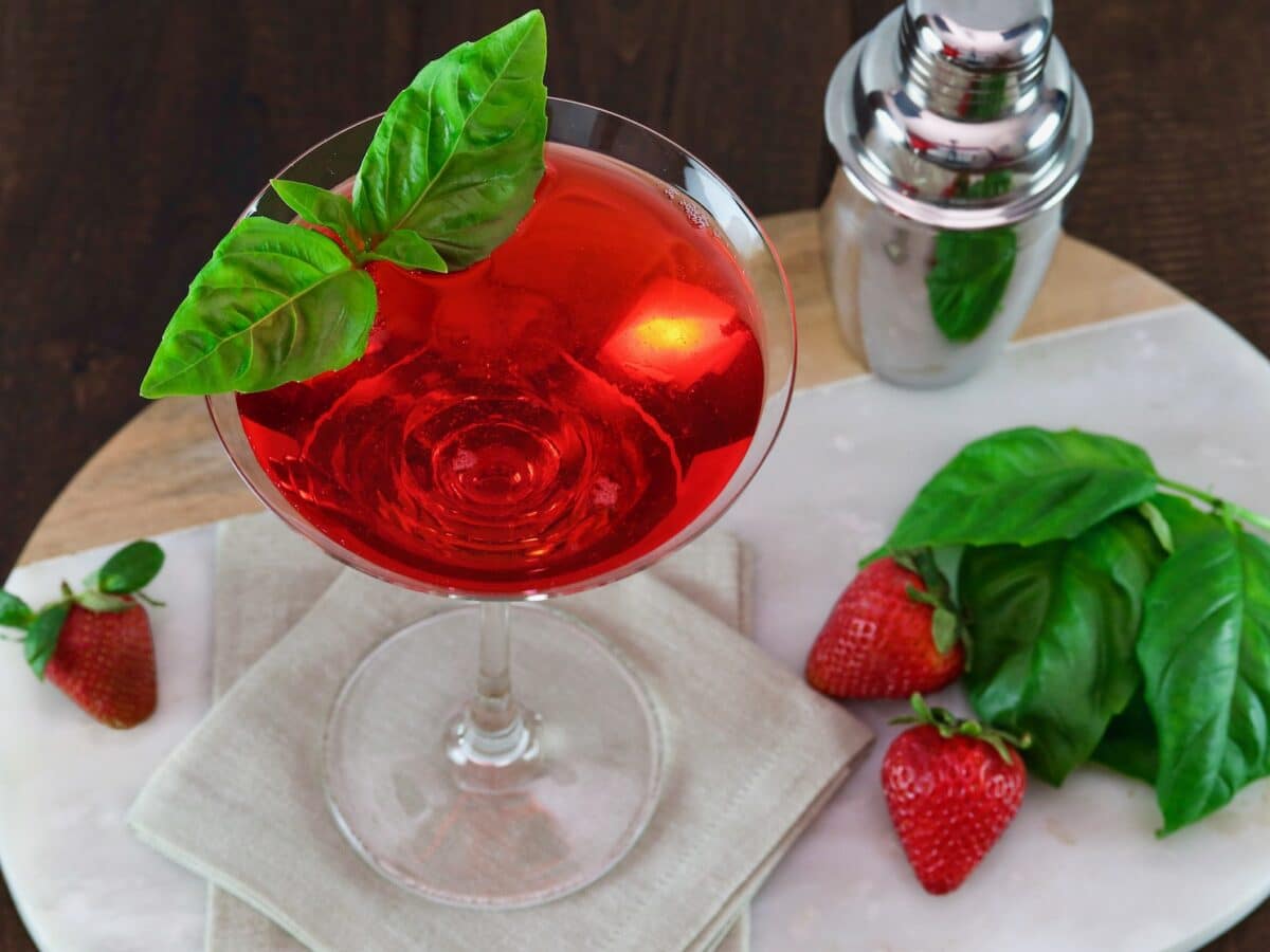 Angled overhead horizontal shot of a martini glass filled with a red strawberry basil martini cocktail, garnished with a sprig of basil. A steel cocktail shaker sits off to the right side next to fresh basil and strawberries.