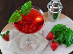 Angled overhead horizontal shot of a martini glass filled with a red strawberry basil martini cocktail, garnished with a sprig of basil. A steel cocktail shaker sits off to the right side next to fresh basil and strawberries.