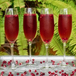 Vertical shot of four champagne flutes filled with a bright red pomegranate cocktail. Pomegranate arils are floating in the cocktails and scattered around the table.