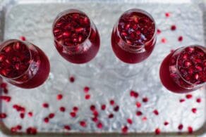 Overhead horizontal shot of four champagne flutes filled with a bright red pomegranate cocktail. Pomegranate arils are floating in the cocktails and scattered around the table.