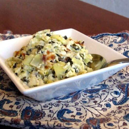 Spinach Artichoke Dip served on a square-shaped bowl with a spoon and on top of a table clothe