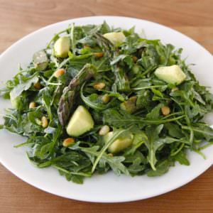 A white plate full of Roasted Asparagus, Avocado and Arugula Salad on topped of a table