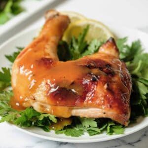 Horizontal shot - whole roast chicken leg topped with honey garlic sauce on a bed of parsley garnish on white plate, platter of chicken in background, on a marble countertop.