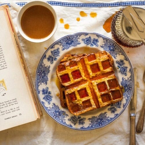 Close-up of two Bread and Butter Waffles soaked on syrup served on a fancy plate with a cup of coffee, syrup glass, and cook book on the side