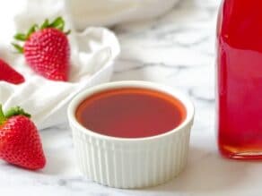 Horizontal Crop - Homemade Strawberry Syrup in a white bowl on a white marble background with fresh strawberries and a white towel, partial view of glass bottle filled with strawberry syrup on the side.