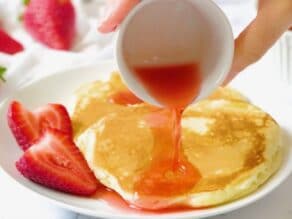 Horizontal crop - hand holding small dosh of strawberry syrup, ouring over homemade pancakes garnished with fresh strawberries on a white plate.
