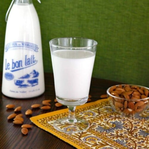 A horizontal image featuring a glass of creamy Almond Milk, with a bowl overflowing with fresh almonds on the left and a bottle of milk on the right.