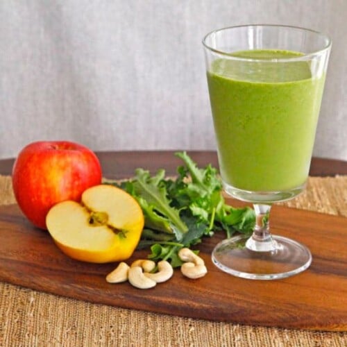 Glass filled with Cashew Apple Green Smoothie surrounded with apples, cashews, and kale