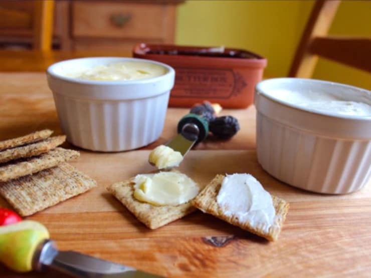 Two crackers spread with homemade butter on a wooden cutting board with two dishes of butter behind - one light colored, one yellow. Knife to spread.