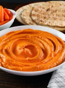 Horizontal shot - white bowl of red pepper hummus with gold spoon, cloth napkin on side. Pita and dish of carrots in background.
