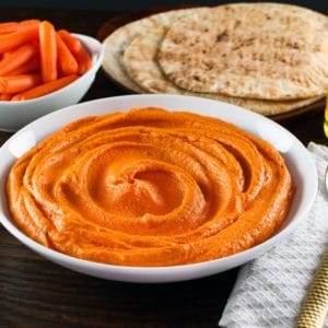 Horizontal shot - white bowl of red pepper hummus with gold spoon, cloth napkin on side. Pita and dish of carrots in background.