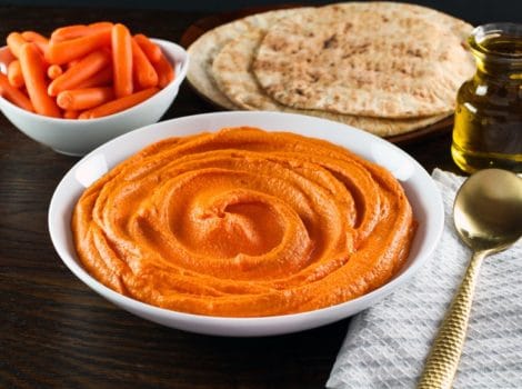 Horizontal shot - white bowl of red pepper hummus with gold spoon, cloth napkin on side. Pita and dish of carrots in background.