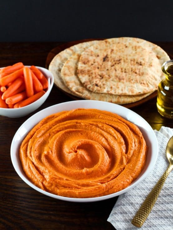 Vertical shot - shallow white bowl of creamy red pepper hummus with gold spoon, cloth napkin on side. Carafe of olive oil, pita and dish of carrots in background.