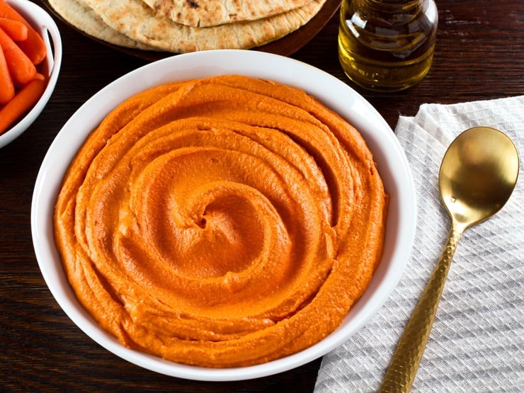 Overhead shot - creamy luscious roasted red pepper hummus in a white dish with gold spoon, cloth napkin on side. Carafe of olive oil, pita and dish of carrots in background.