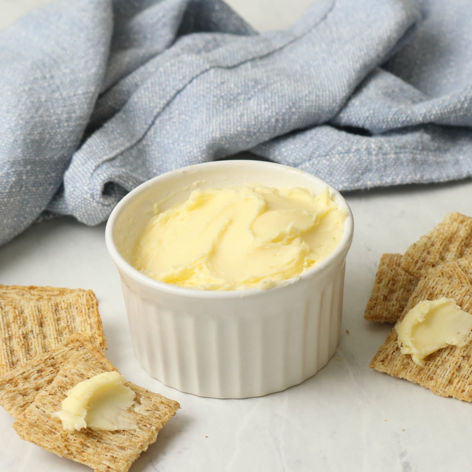 A dish of homemade butter on a marble surface, crackers spread with butter. Blue towel in background.