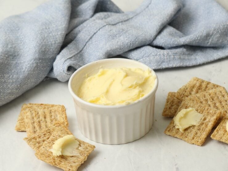 A dish of homemade butter on a marble surface, crackers spread with butter. Blue towel in background.