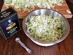 Bowl of shredded cabbage on wooden table with teaspoon and box of salt, cutting board with shredded cabbage in background.