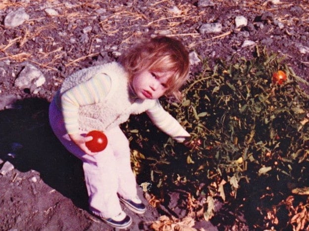 Tori Avey as a toddler in her grandfather's garden holding a tomato, leaning over a tomato plant.