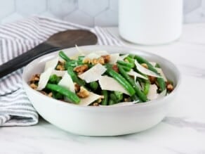 Green bean salad with walnuts and parmesan as garnish on a medium-sized white bowl in foreground with a wooden spoon. Linen napkin and canister in background.