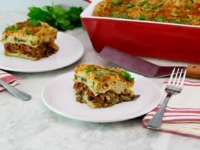 A square slice of vegan moussaka on a small white plate in foreground with fork and linen napkin. A red, rectangular baking dish and a stainless steel serving shovel on the background with another serving of a sliced vegan moussaka.