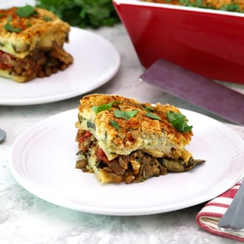 A square slice of vegan moussaka on a small white plate in foreground with fork and linen napkin. A red, rectangular baking dish and a stainless steel serving shovel on the background with another serving of a sliced vegan moussaka.