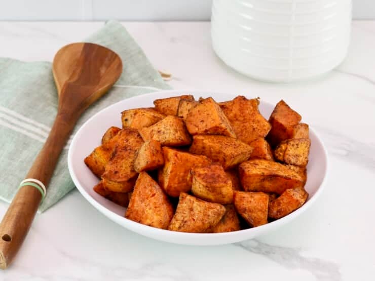 A serving of curry roasted sweet potatoes on a white bowl in foreground with a wooden spoon and linen napkin on the left side. White canister in the background.
