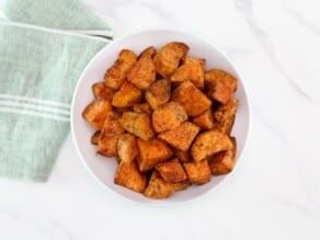 A serving of curry roasted sweet potatoes on a white bowl in foreground with a linen napkin on the left side.