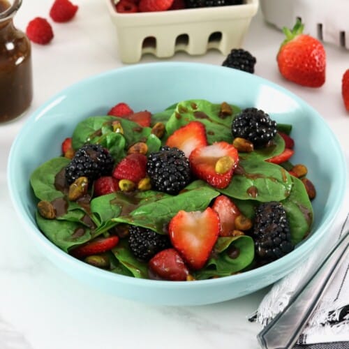 Strawberry spinach salad on a light blue plate topped with maple balsamic dressing in foreground with fork, and gray and white stripe linen napkin. Garnish of strawberries, blackberries, and raspberries, a glass carafe with maple balsamic dressing, and a white and cream colored plastic basket containers with lots of strawberries, raspberries, and blackberries, in background.