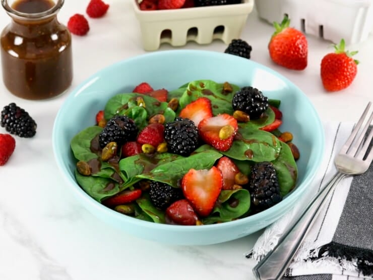 Strawberry spinach salad on a light blue plate topped with maple balsamic dressing in foreground with fork, and gray and white stripe linen napkin. Garnish of strawberries, blackberries, and raspberries, a glass carafe with maple balsamic dressing, and a white and cream colored plastic basket containers with lots of strawberries, raspberries, and blackberries, in background.