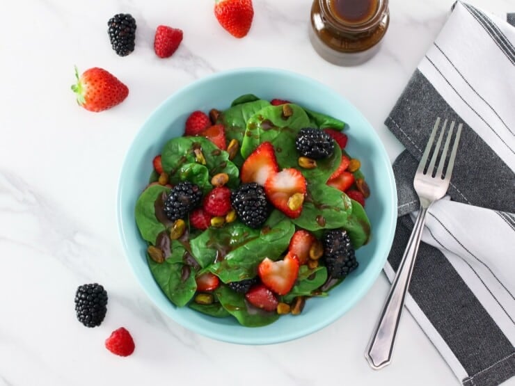 Overhead shot - spinach strawberry salad on a light blue plate topped with maple balsamic dressing, strawberries, blackberries, raspberries and pistachios in foreground. Table set with fork and gray and white stripe linen napkin. Garnish of four small strawberries and two blackberries, and a small glass carafe bottle with maple balsamic dressing, in background.