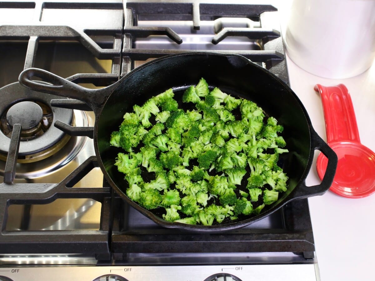 Broccoli florets in a large skillet over the top of cooking range. A red teaspoon on the side, and canister in background.