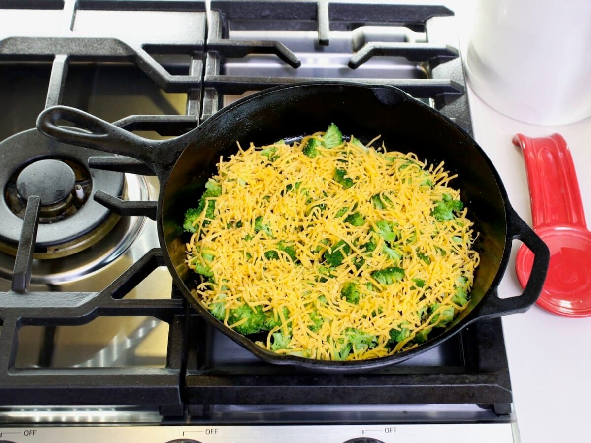 Broccoli florets topped with an even layer of grated cheese in a large skillet over the top of cooking range. A red teaspoon on the side, and canister in background.