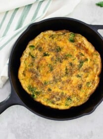 A whole circular broccoli cheese frittata in a large skillet in foreground. Garnish of fresh dill on the side, linen napkin in background.