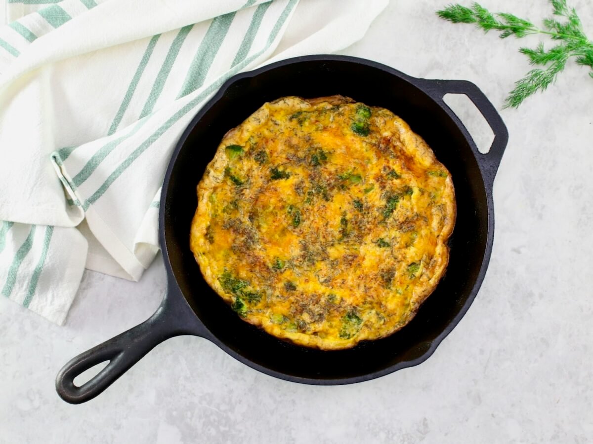 A whole circular broccoli cheese frittata in a large skillet in foreground. Garnish of fresh dill on the side, linen napkin in background.