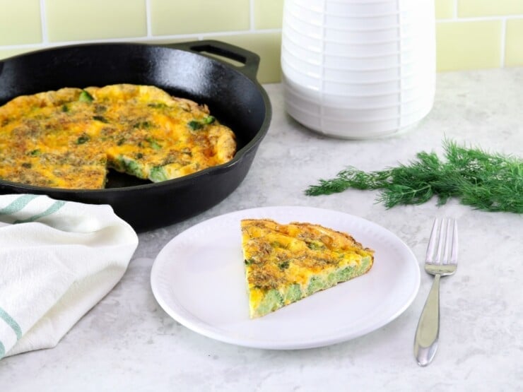 A triangular slice of broccoli cheese frittata on a small white plate in foreground with fork, larger frittata in skillet in background. Garnish of fresh dill on the side, linen napkin and canister in background.